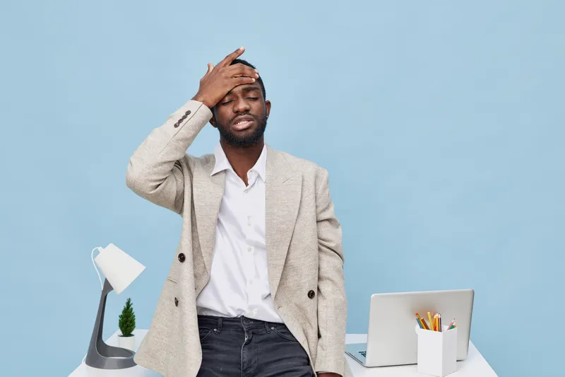 Frustrated man in business casual outfit sitting at desk with laptop and office supplies, holding his head in stress or disappointment, isolated on plain light blue background. People concept