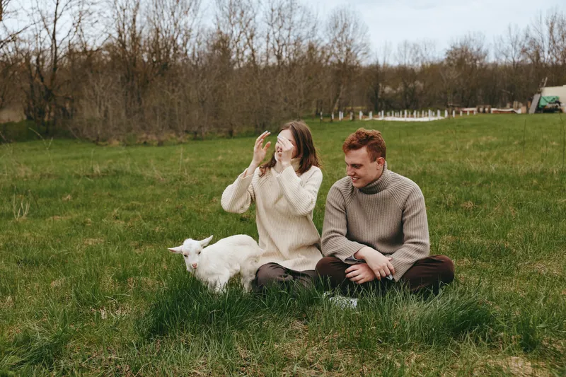 Casual couple sitting in a grassy field with a playful goat kid nearby, enjoying a relaxed outdoor moment, natural light, candid smiles, simple countryside vibe