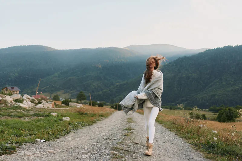 A woman in casual outdoor attire stands on a gravel path in a mountain valley, looking toward the distance. A calm, reflective moment in nature and lifestyle.