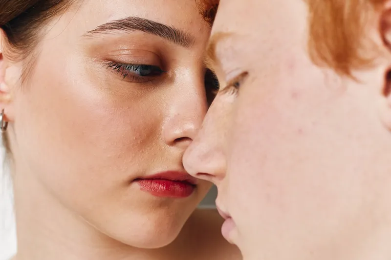 Closeup portrait of young woman and man face to face with eyes closed for intimacy, connection, love, and emotion in soft natural light. Skin texture, freckles, and makeup visible.