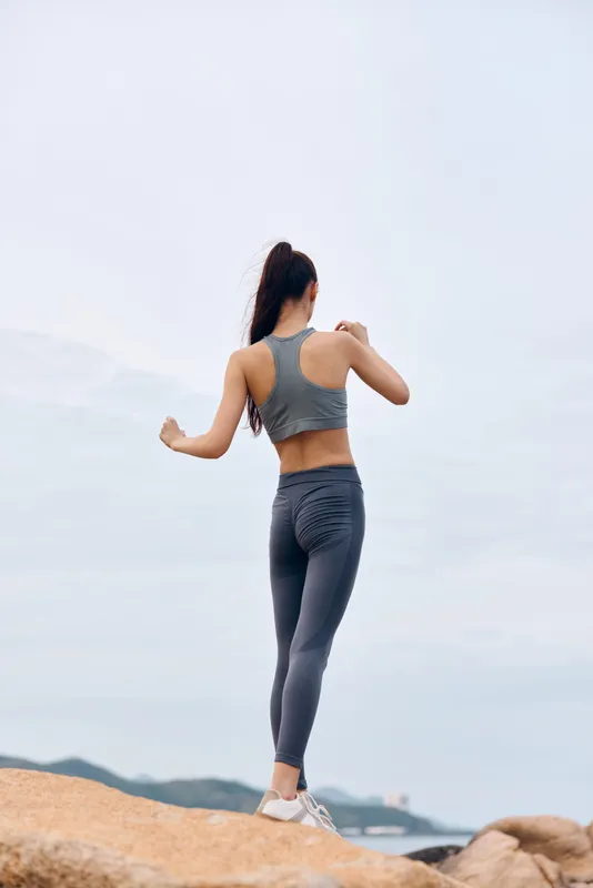 Fit female athlete in gray activewear stretching on a rocky beach, showcasing strength, determination, and a healthy lifestyle against a soft sky background