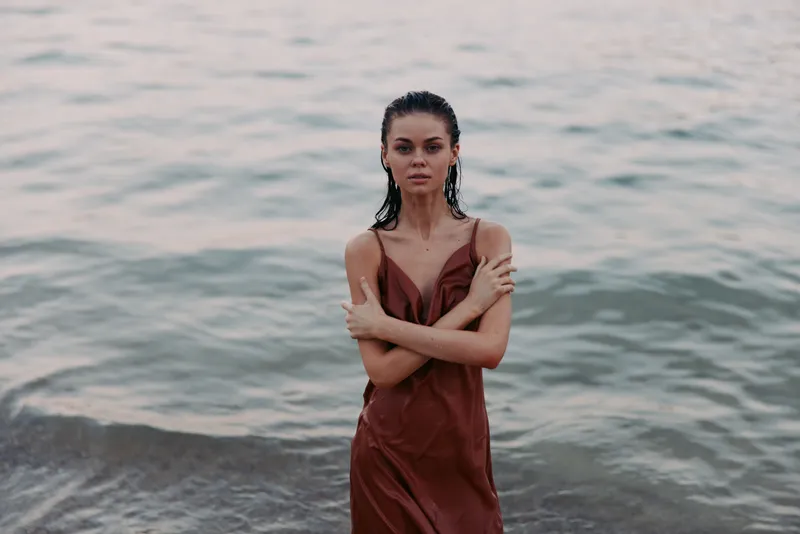 A solitary woman stands in shallow coastal water, wearing a rust red dress, arms crossed, facing the camera with a quiet, confident gaze and a serene, minimalist mood.