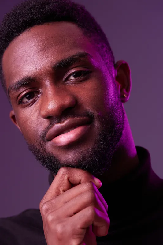 Confident young Black man in a black turtleneck sweater with a thoughtful expression, illuminated by vibrant purple lighting, ideal for fashion and personal growth themes
