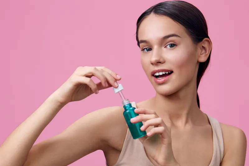 Young woman holding a green serum bottle, smiling joyfully against a pink background, embodying beauty, skincare, and confidence in a fresh, youthful style