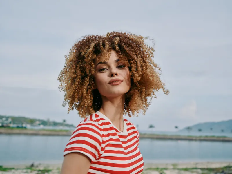 Portrait of a young woman with curly hair wearing a red and white striped tee, standing by a calm coastal scene under soft daylight.