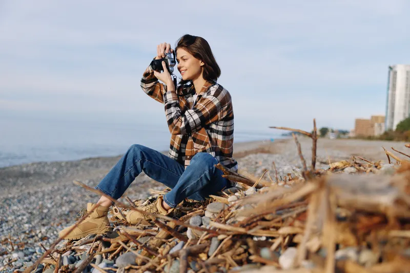 Casual photographer, a woman on a sandy beach with camera, crouching among driftwood and pebbles to capture the ocean and distant skyline at the shoreline.