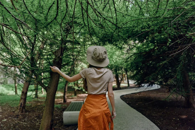 Woman walking on a shaded park path among tall trees, wearing a hat and orange skirt, hand touching trunk beside a bench, nature stroll with serene green canopy.