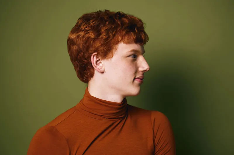 Profile portrait of young redhead man with curly hair wearing brown turtleneck against olive background. Side view, thoughtful expression and casual fashion style in studio lighting.