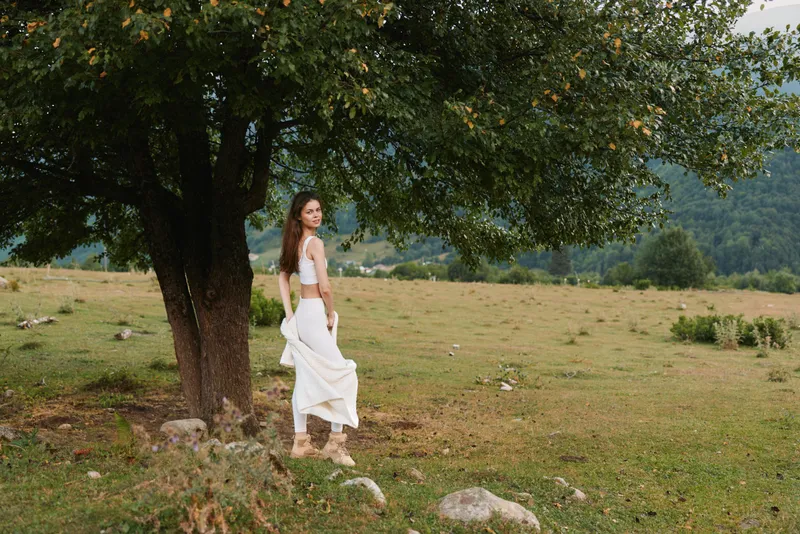 Young woman in a white dress stands beside a large tree in an open field, calm and reflective under soft natural light.