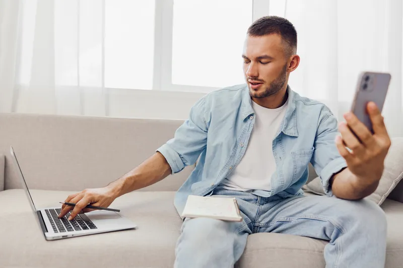 Focused man multitasking with a laptop and smartphone, showcasing productivity and modern lifestyle in a cozy home environment. Perfect for digital nomad themes.