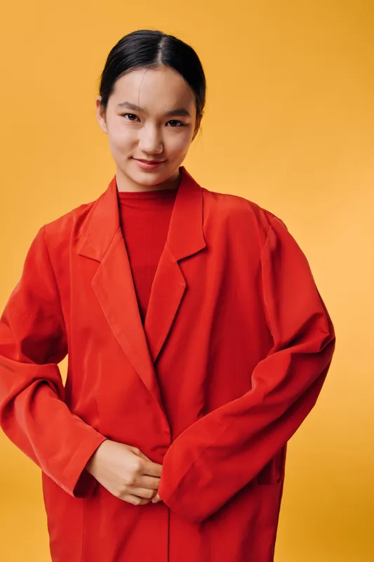 Young woman wearing a red coat standing against orange background, smiling slightly with hands adjusting the front. Studio portrait with bright colors and confident expression.