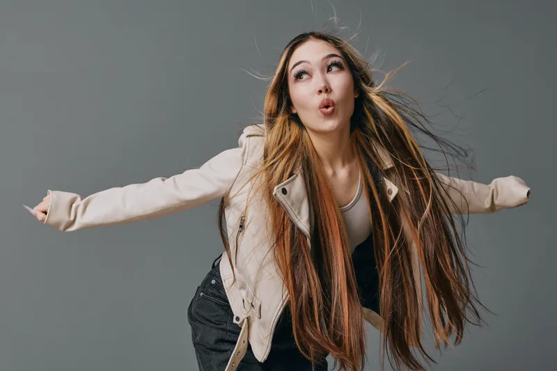 Young woman with long hair wearing a light jacket making a playful face and balancing with arms outstretched on grey background. Casual expression and motion captured in studio.