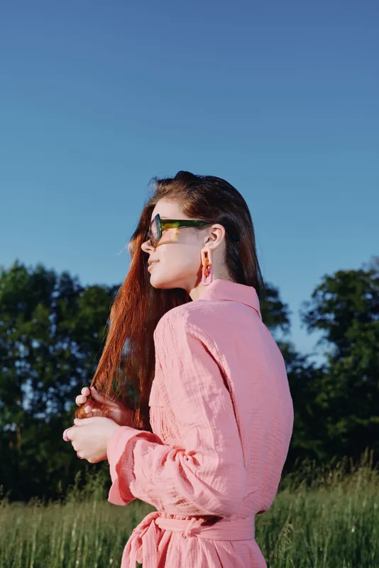 Young woman in a pink dress holding her hair, enjoying a sunny day outdoors in a vibrant green field, conveying a sense of freedom and positivity