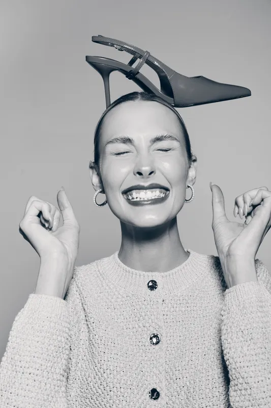 Young white woman with fair skin and closed eyes smiles broadly holding her hands up, balancing high heel shoe on her head. Concept of joy and fashion fun.