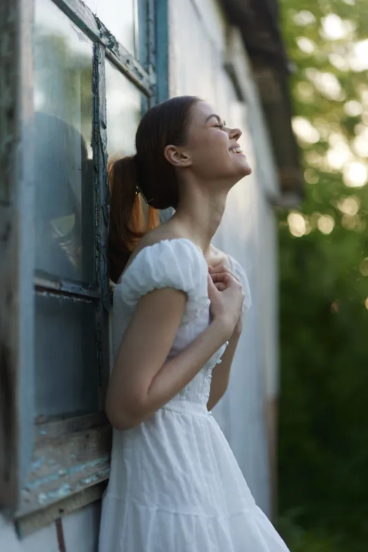 Young woman in a white dress standing by a window, smiling joyfully, surrounded by greenery in a sunny outdoor setting, conveying feelings of happiness and serenity