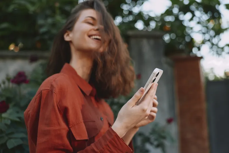 A smiling woman in a red shirt enjoys a sunny outdoor moment, looking at a phone. A candid lifestyle scene showing casual confidence, outdoor leisure and everyday technology use