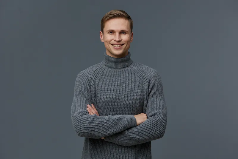 Smiling man wearing cozy gray turtleneck sweater standing against plain neutral background in studio portrait shot. Casual fashion, happy mood, confident expression, modern style, indoor setting, wint
