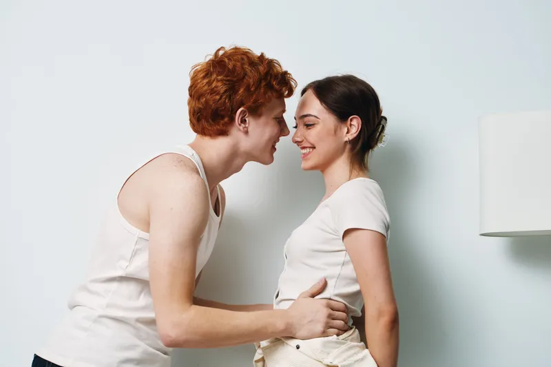 Young couple standing close to each other indoors smiling and looking into each other eyes showing affection and happiness with casual clothes against white wall background.