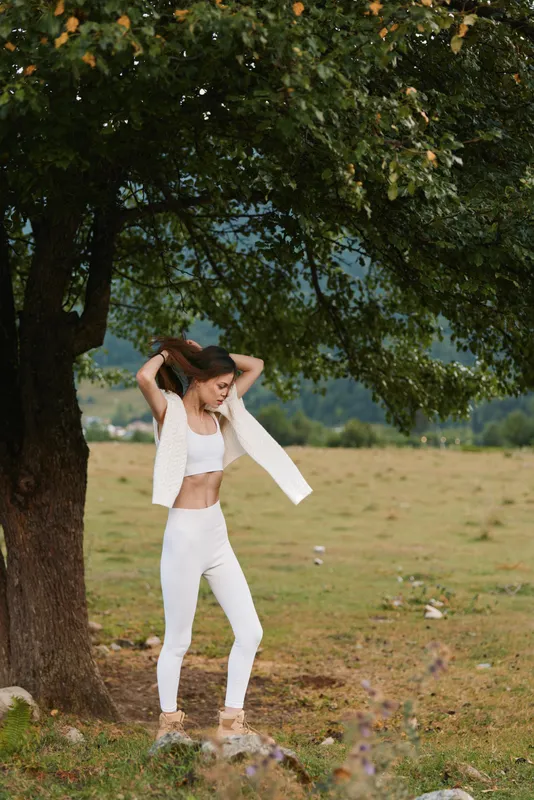 Young woman in white outfit stands under tree in nature, adjusts hair, enjoys outdoor lifestyle, casual fashion, peaceful countryside background.