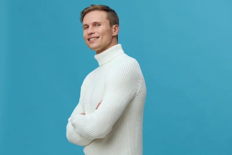 Smiling man in white turtleneck sweater posing confidently against a bright blue background. Casual clothing, friendly expression, natural hairstyle, studio shot, cheerful mood, people lifestyle conce