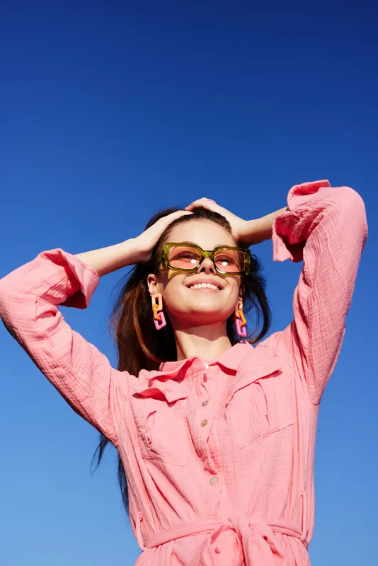 Young woman in stylish pink outfit with colorful sunglasses smiles joyfully against a clear blue sky, embodying summer vibes and cheerful fashion
