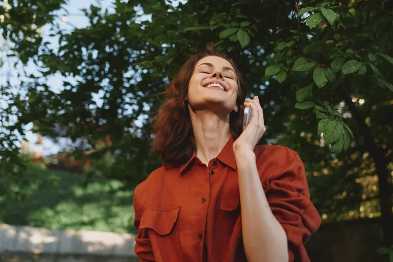 A smiling woman outdoors in a red shirt listens to music with headphones, enjoying sunny nature, a casual lifestyle moment that radiates happiness
