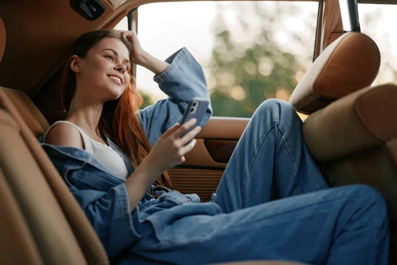 Young woman in casual denim outfit enjoying her phone in the back seat of a car, showcasing joy and relaxation in a sunny outdoor setting.
