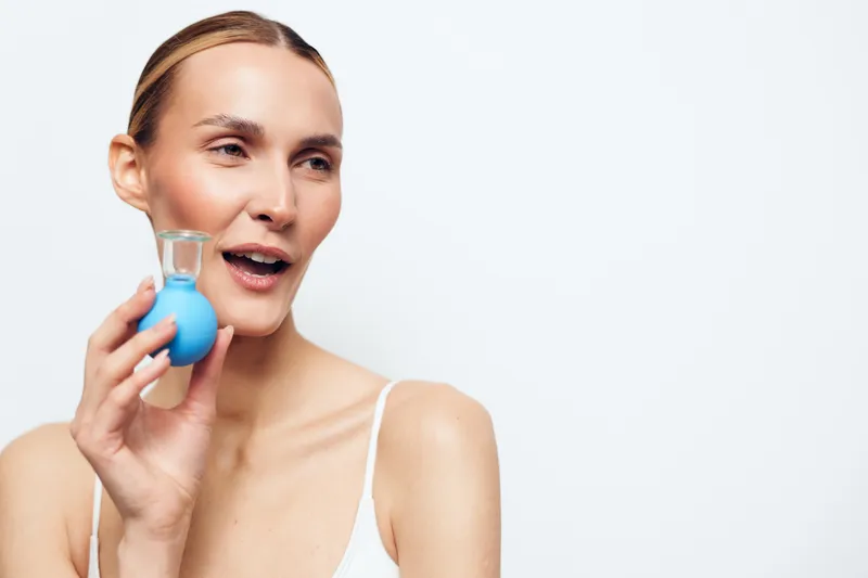 A smiling woman holds a small blue bottle near her face, showcasing a beauty product for skincare or fragrance in a bright studio, casual outfit, soft lighting, minimal background, cheerful mood,