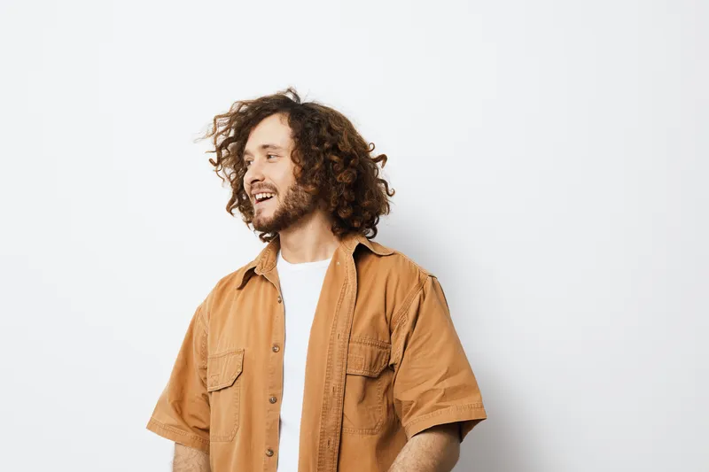 Young man with curly hair wearing a casual brown shirt, smiling joyfully against a light background, embodying positivity and confidence