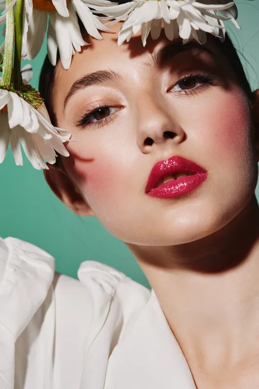 Closeup portrait of young woman with bright lipstick and floral crown on head poses on green background. The image focuses on beauty, fashion, and creative makeup styling.