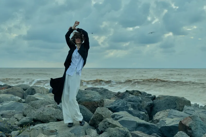 Fashion, model on rocky coastline, windblown coat and dress, dynamic pose by the sea, dramatic clouds, outdoor editorial in natural light, coastal fashion, movement and energy, ocean backdrop