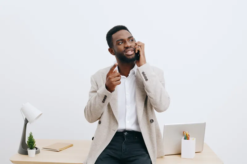 Confident young smiling man with dark skin, curly hair, and a beard, wearing a beige blazer and white shirt, talking on mobile phone, in modern office setting with white background. People lifestyle c