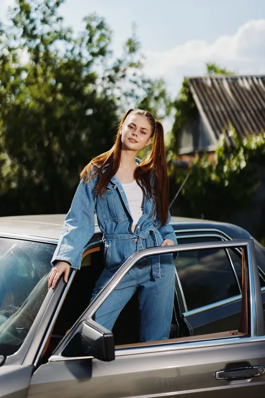 Stylish young woman in denim jumpsuit, posing confidently by vintage car in sunny outdoor setting, showcasing a carefree and trendy vibe.