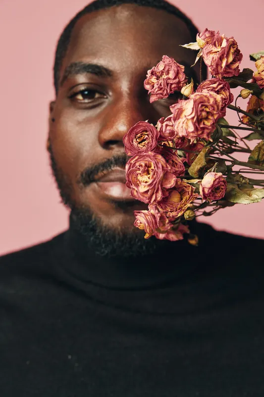 African man with beard and black turtleneck holding dried pink roses close to face on pink background creative conceptual visual style portrait with natural expression and detail