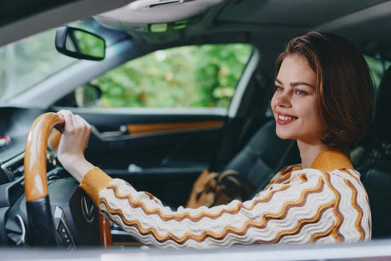Portrait of a smiling woman driving a car, gripping a wooden steering wheel, with a cozy sweater and warm interior styling.
