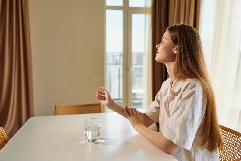 woman table glass window sunlight portrait morning home: Young woman sits at a white table by a window bathed in sunlight, holding her arm and gazing outside with a glass of water, relaxed domestic