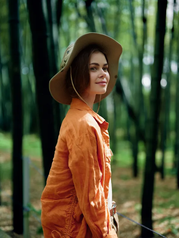 Woman portrait in hat and orange jacket standing in bamboo forest outdoors, smiling gently at camera with natural light, casual style and serene green background for lifestyle photography.