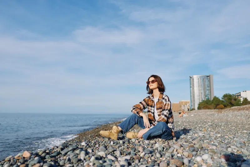 Beach, sea, woman sitting on pebbled coast in casual plaid shirt and sunglasses, enjoying outdoor relaxation by the water, with distant city skyline and clear blue sky.