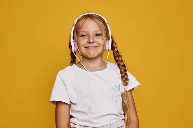 Smiling young girl with braids wearing headphones and a white t-shirt stands against an orange background. Concept of music, happiness, childhood, and technology.
