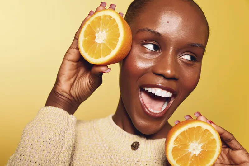 Happy dark skinned African woman with short hair holds two halves of fresh orange fruit near face on yellow background. Concept of healthy lifestyle, vitamin, and natural beauty.