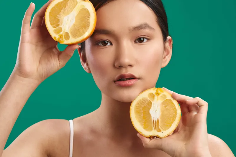 asian woman holds fresh orange halves against a vibrant green backdrop, showcasing healthful produce and a lively studio mood. Her expression is calm and confident, highlighting natural beauty and