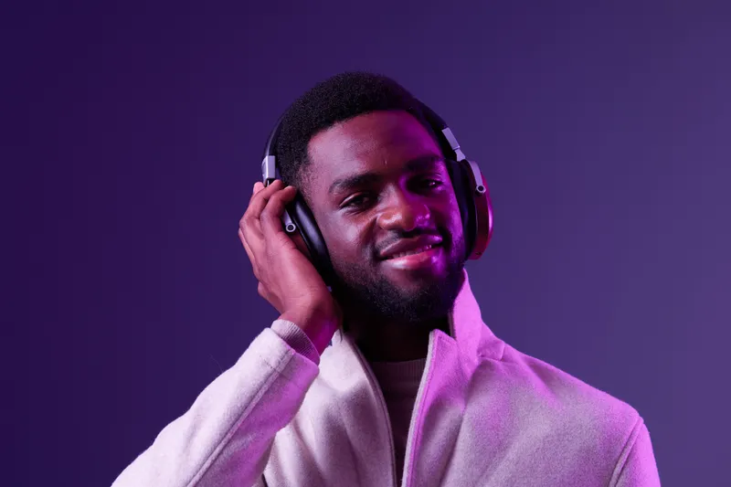 Stylish young Black man enjoying music with headphones, smiling happily against a vibrant purple background, perfect for lifestyle and technology themes