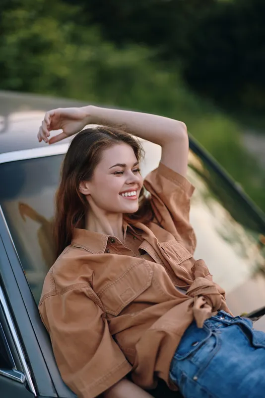 Happy young woman in casual brown shirt resting on a vintage car, enjoying a sunny day outdoors with green background.