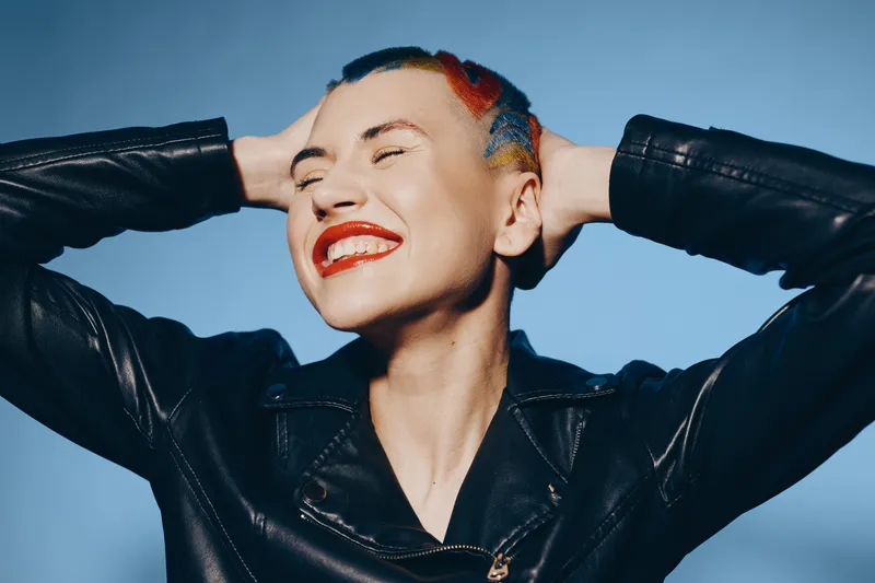 Smiling woman with colorful short hair wearing a black leather jacket, posing confidently against a clear blue sky background with bright natural light.