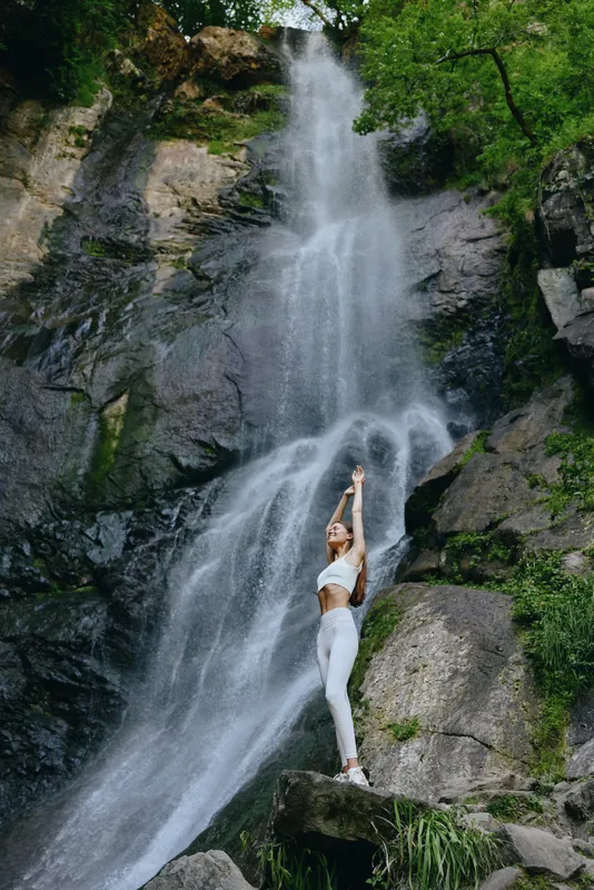 woman stretching near waterfall in white activewear stands on rock surrounded by lush green forest creating a peaceful and refreshing outdoor scene.