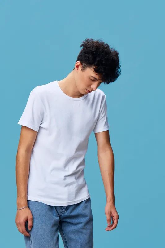 Young man with curly hair wearing white t-shirt and blue jeans stands looking down thoughtfully against bright blue background in casual style.