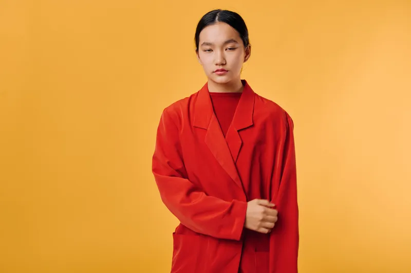 Young woman with black hair wears oversized red coat standing against plain orange background in studio. Casual style, serious expression, minimalistic fashion portrait with clean composition and