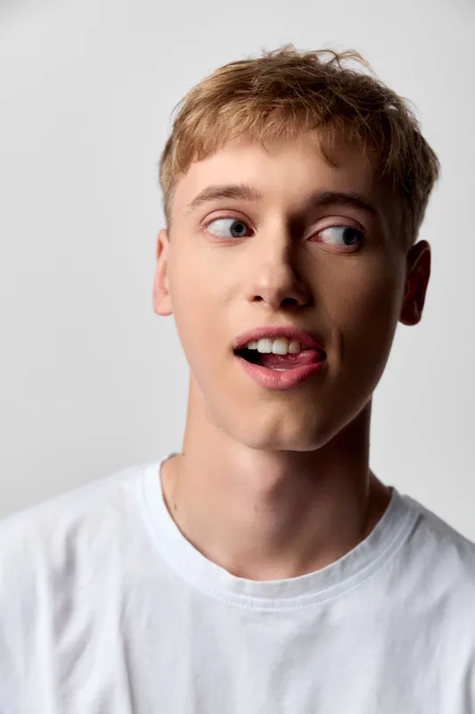 Close up of a youthful man in a white tee, sticking his tongue out playfully, wide eyes, casual studio lighting and plain background, expressive, modern mood, lifestyle photography.