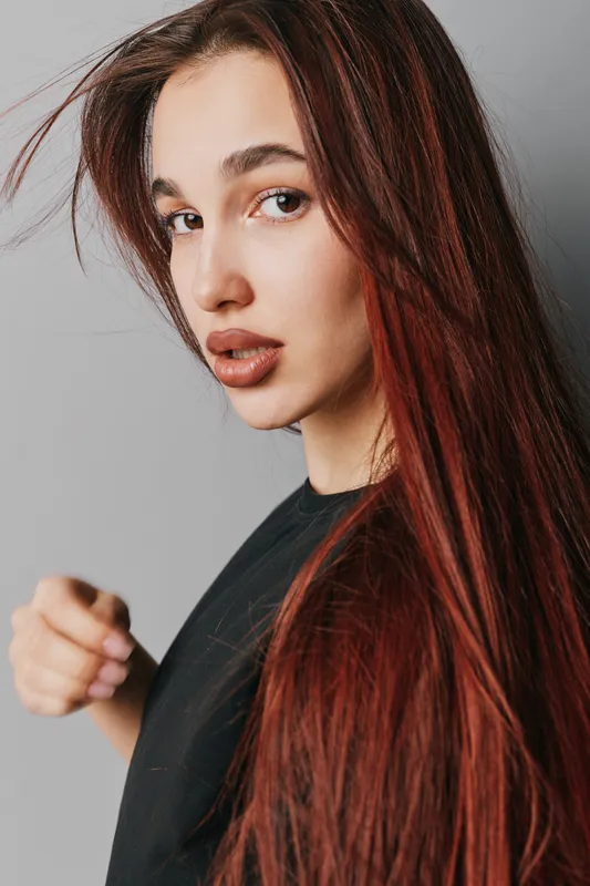 Young woman with long red hair and natural makeup posing in black shirt against gray background. Portrait of confident female with loose hair and serious expression in studio lighting.