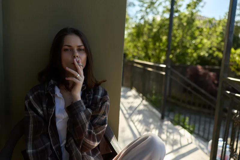 Woman lifestyle outdoor balcony cigarette smoking casual portrait relaxed calm mood on sunny urban terrace, depicting a candid personal moment